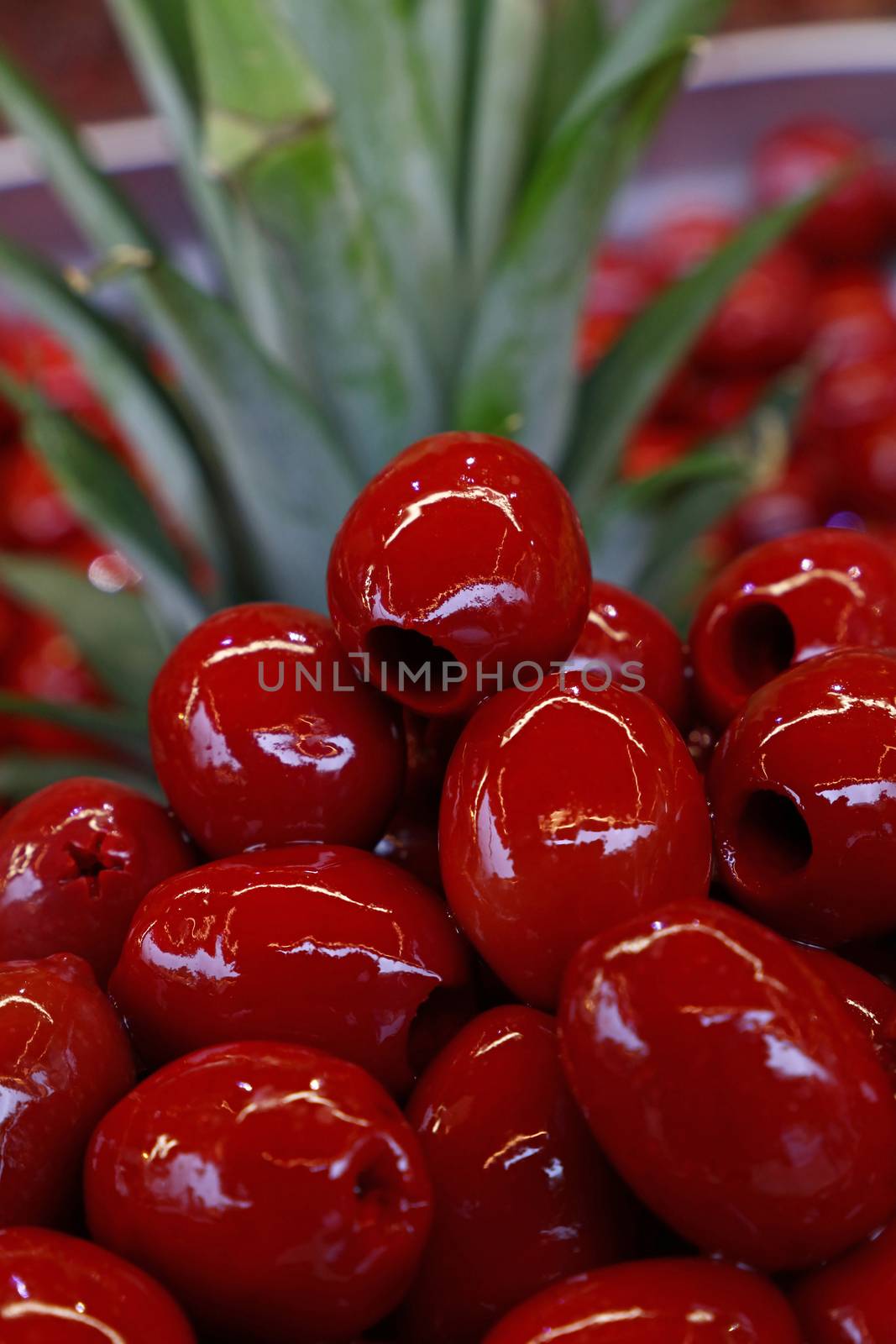Red pitted glossy Italian Cerignola olives in oil close up over green pineapple leaves, retail market stall display, low angle view