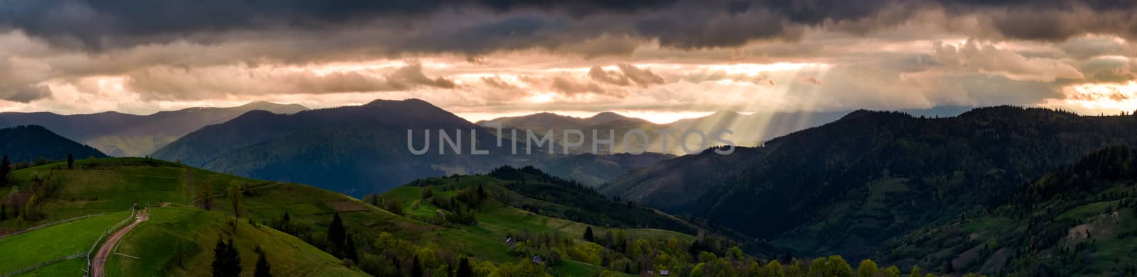 panorama of mountainous countryside at sunset. country road through rolling hills in to the distance. heavy clouds over the ridge. beams of lite from the heaven. beautiful and mysterious landscape