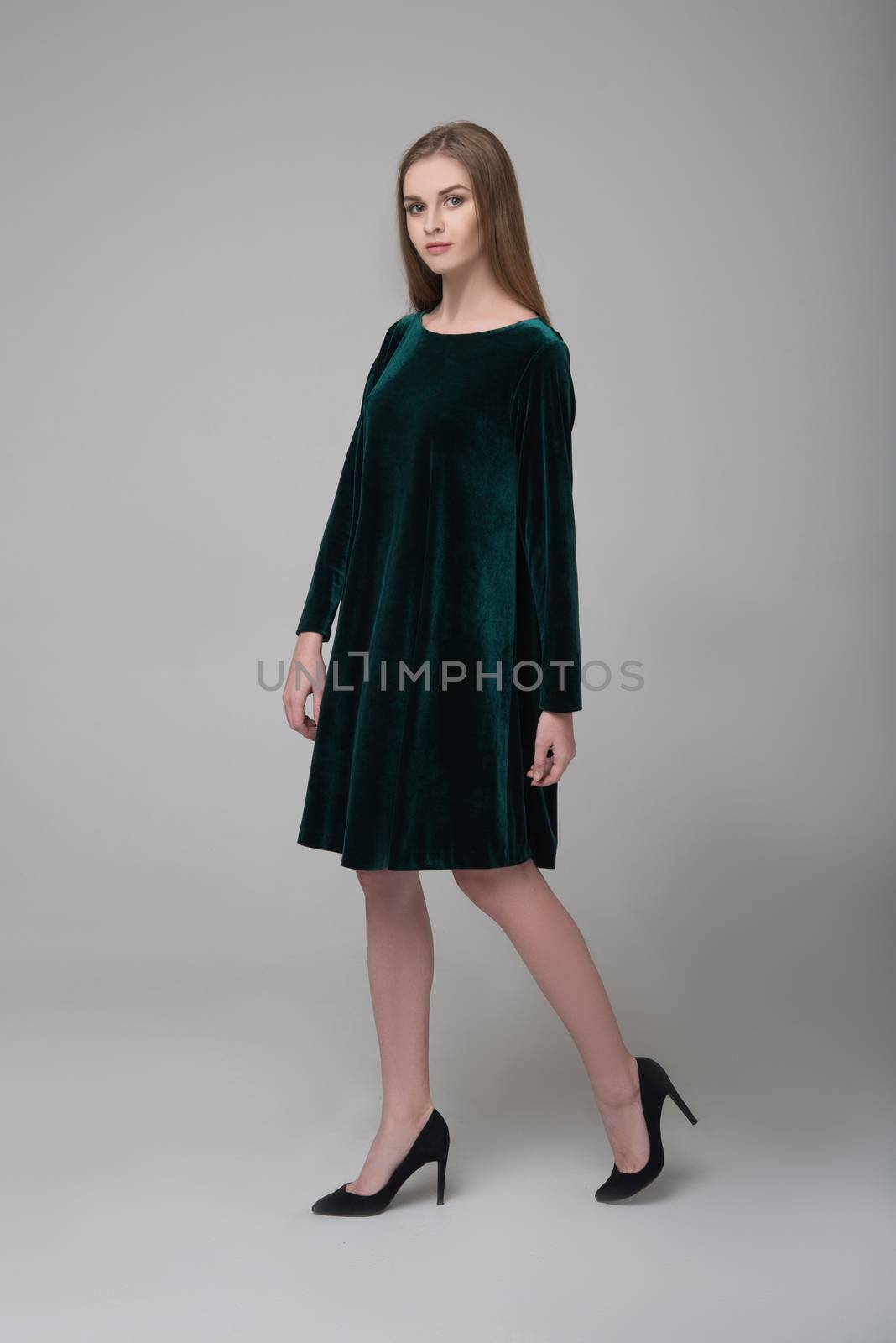 Young beautiful long-haired female model poses in dark green short dress on grey background