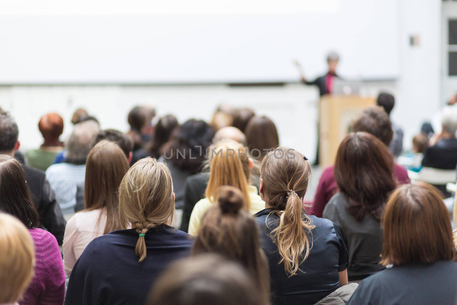 Female speaker giving presentation in lecture hall at university workshop. Audience in conference hall. Rear view of unrecognized participant in audience. Scientific conference event.