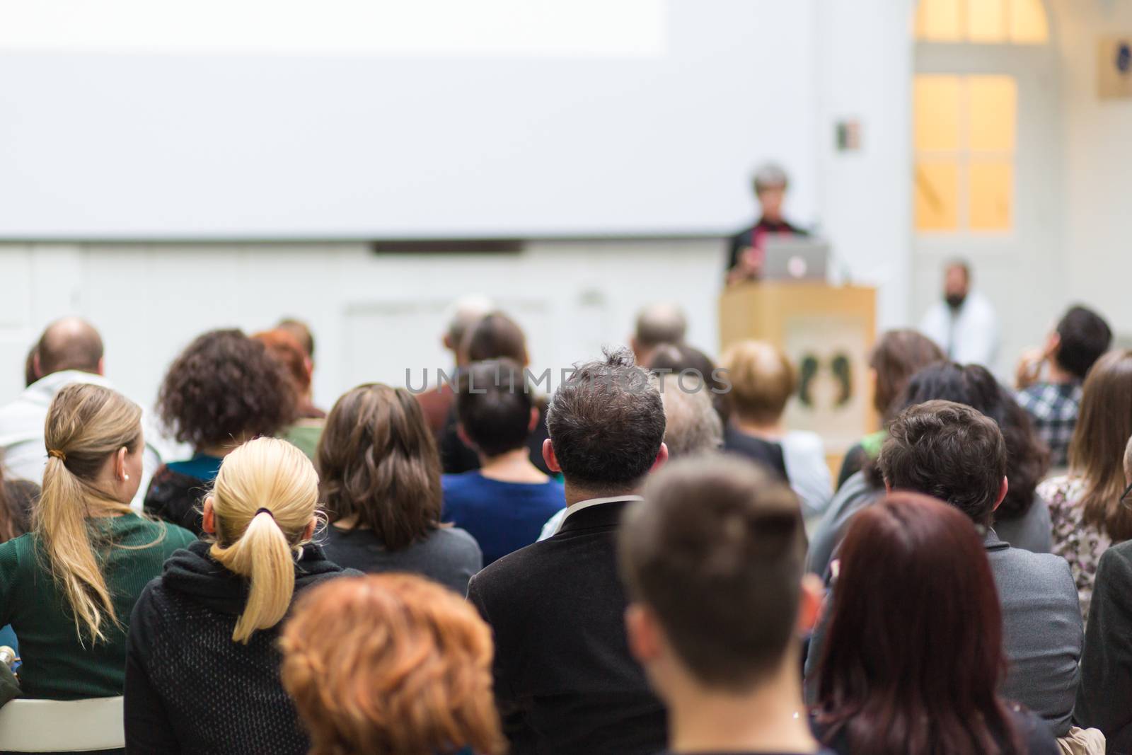 Female speaker giving presentation in lecture hall at university workshop. Audience in conference hall. Rear view of unrecognized participant in audience. Scientific conference event.