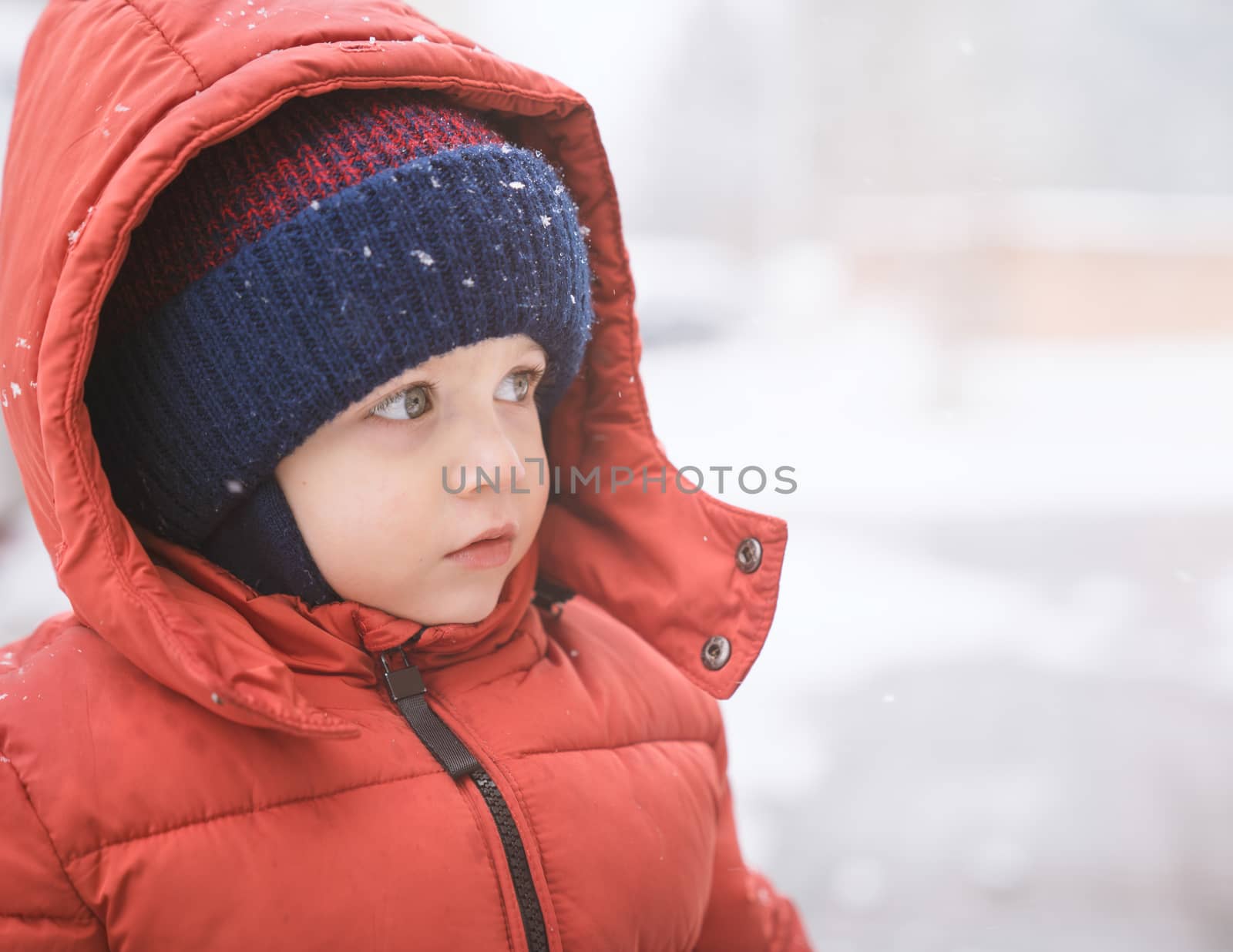 Baby boy while it snows looks towards the emptiness, covered with red winter jacket and woolen hat, close-up.