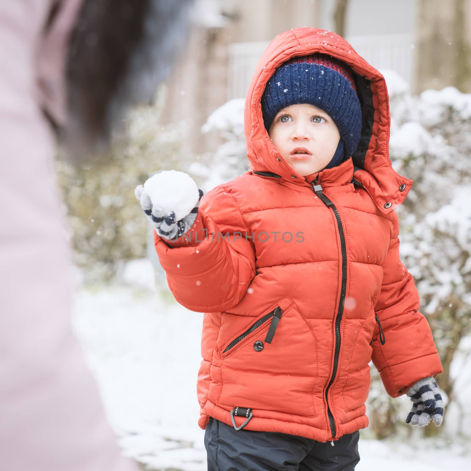 Child while it snows looks towards the mother with a snowball in his hands