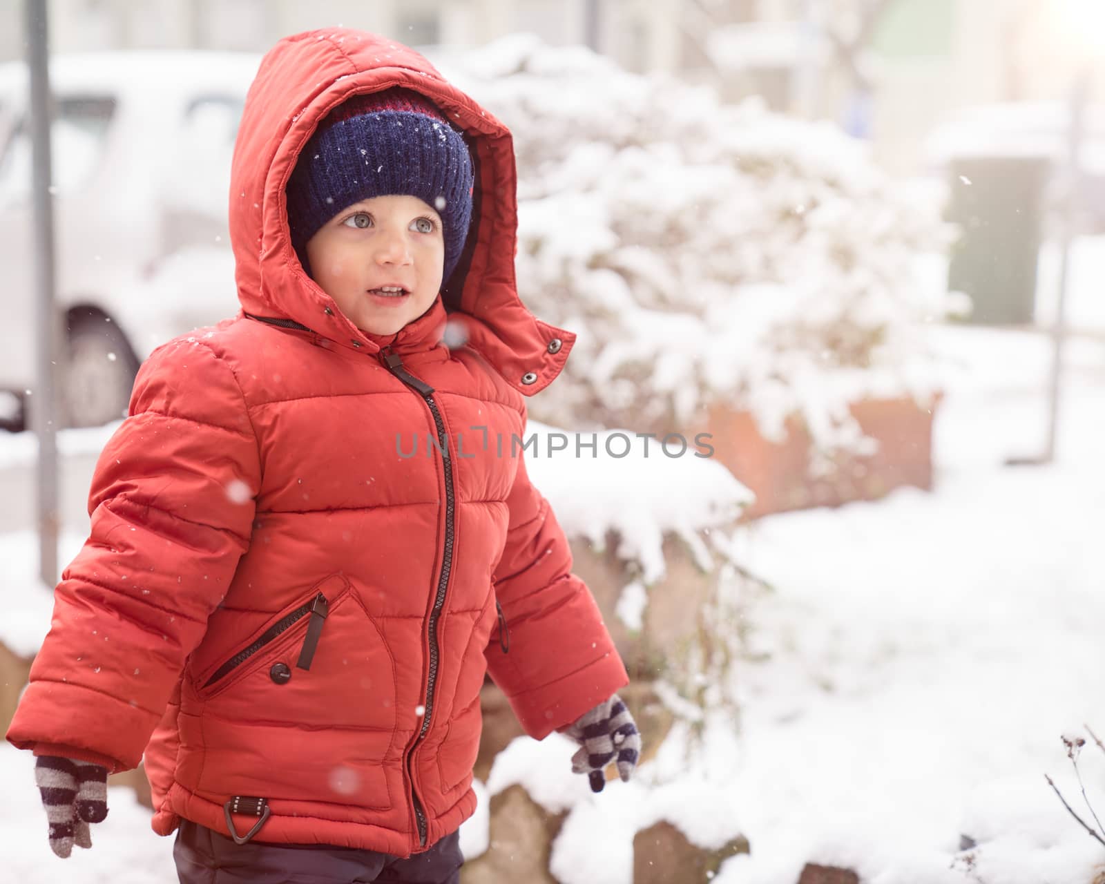 Kid boy while it snows looks towards the emptiness, covered with red winter jacket and woolen hat.