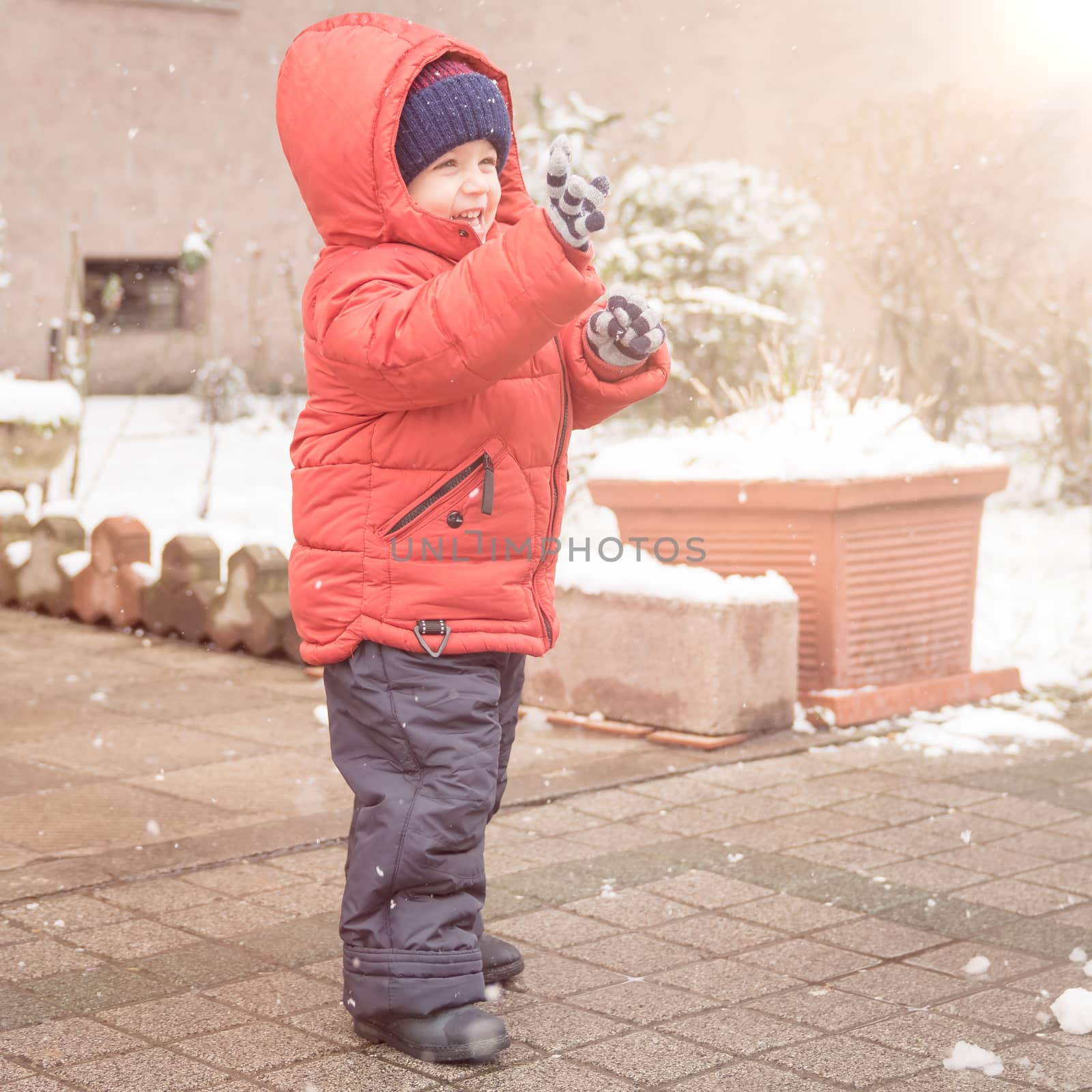 Infant boy smiles cheerfully while it's snowing,dressed in a red winter jacket and a woolen hat.