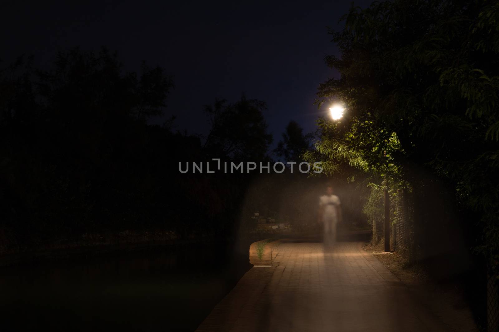 Double exposure night scene of person walking dark street illuminated with streetlights. The receding male silhouettes on the road in the park. Human figure in motion blur going along the city river by MSharova