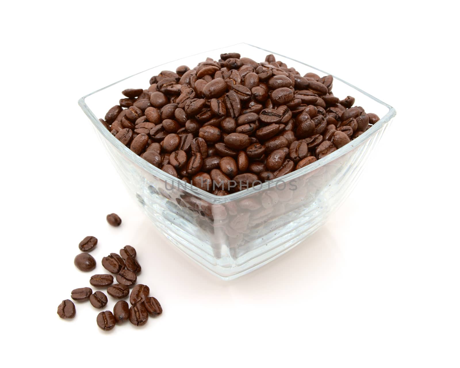 Glass dish filled with roasted coffee beans with some spilled beside on a white background