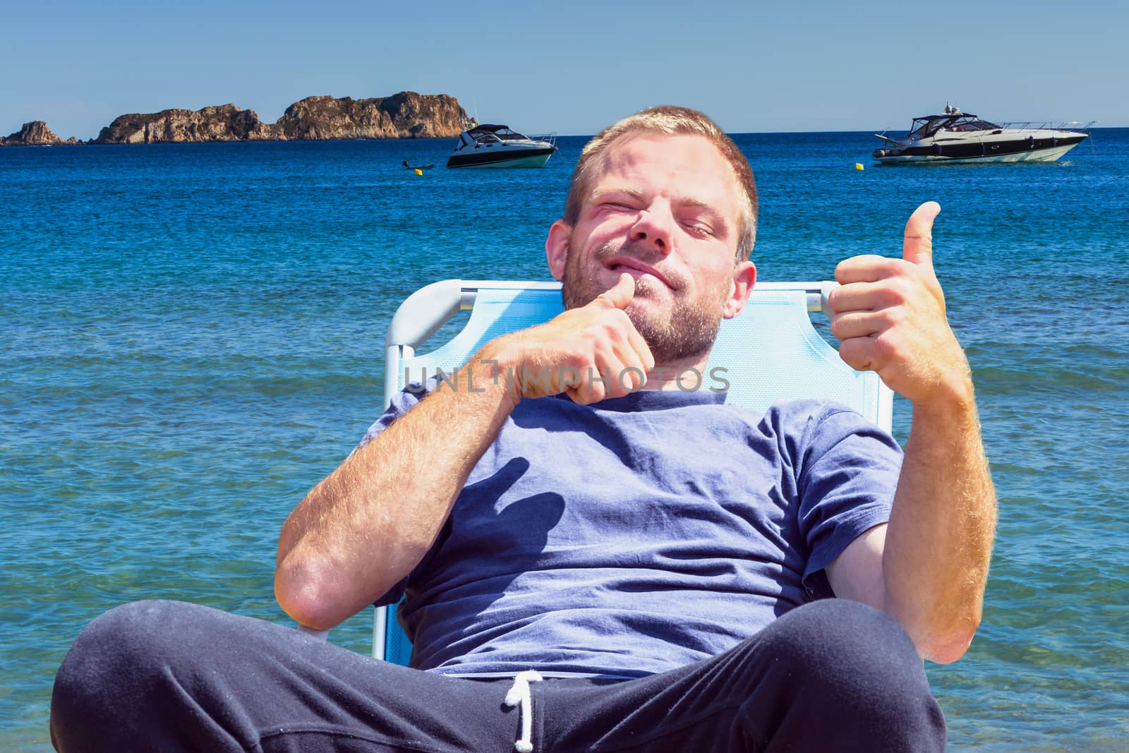 Young man sitting in the deck and holding his thumbs up. In the background the sea and two sports boats.