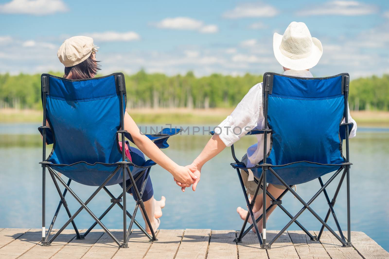 couple in love sitting on chairs at pier near lake and holding hands