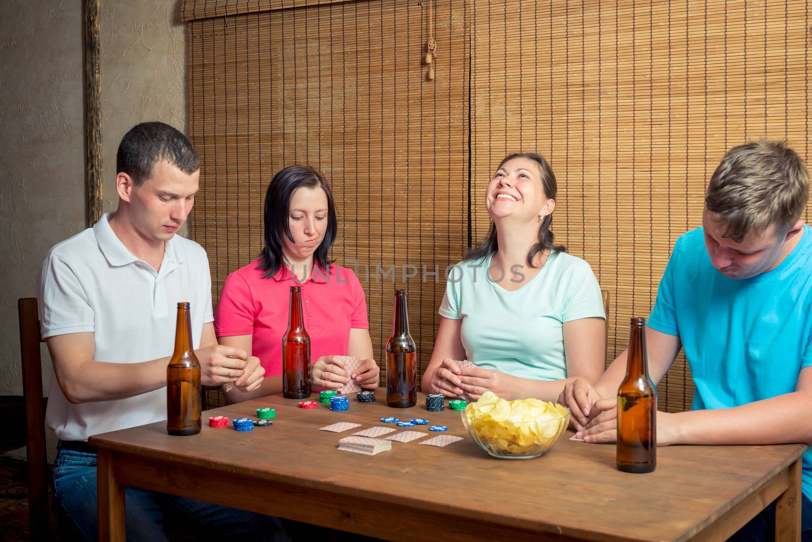 young friends playing cards sitting at a table indoors, drinking beer
