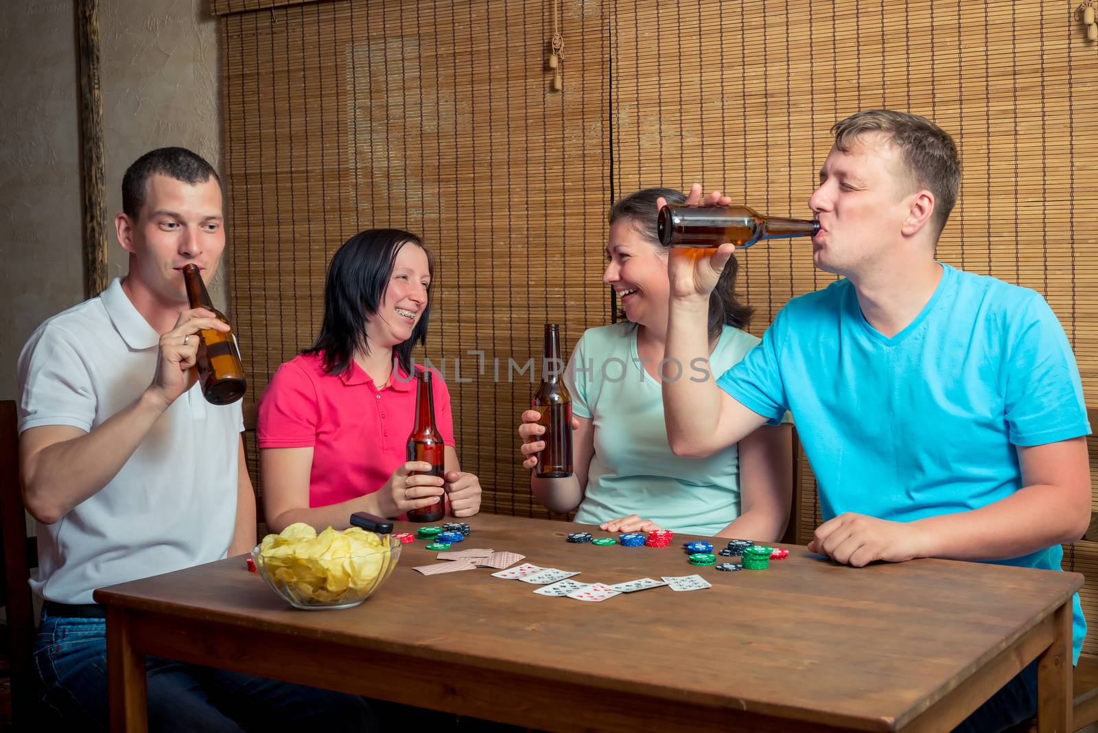 a company of friends drinking beer and playing cards sitting at a table indoors