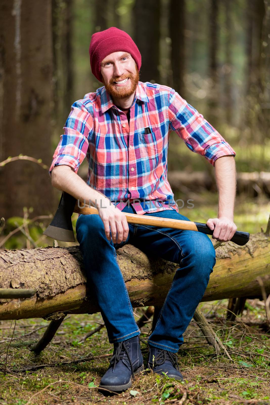 a smiling bearded lumberjack in a hat and shirt with an ax sits on a log in the woods
