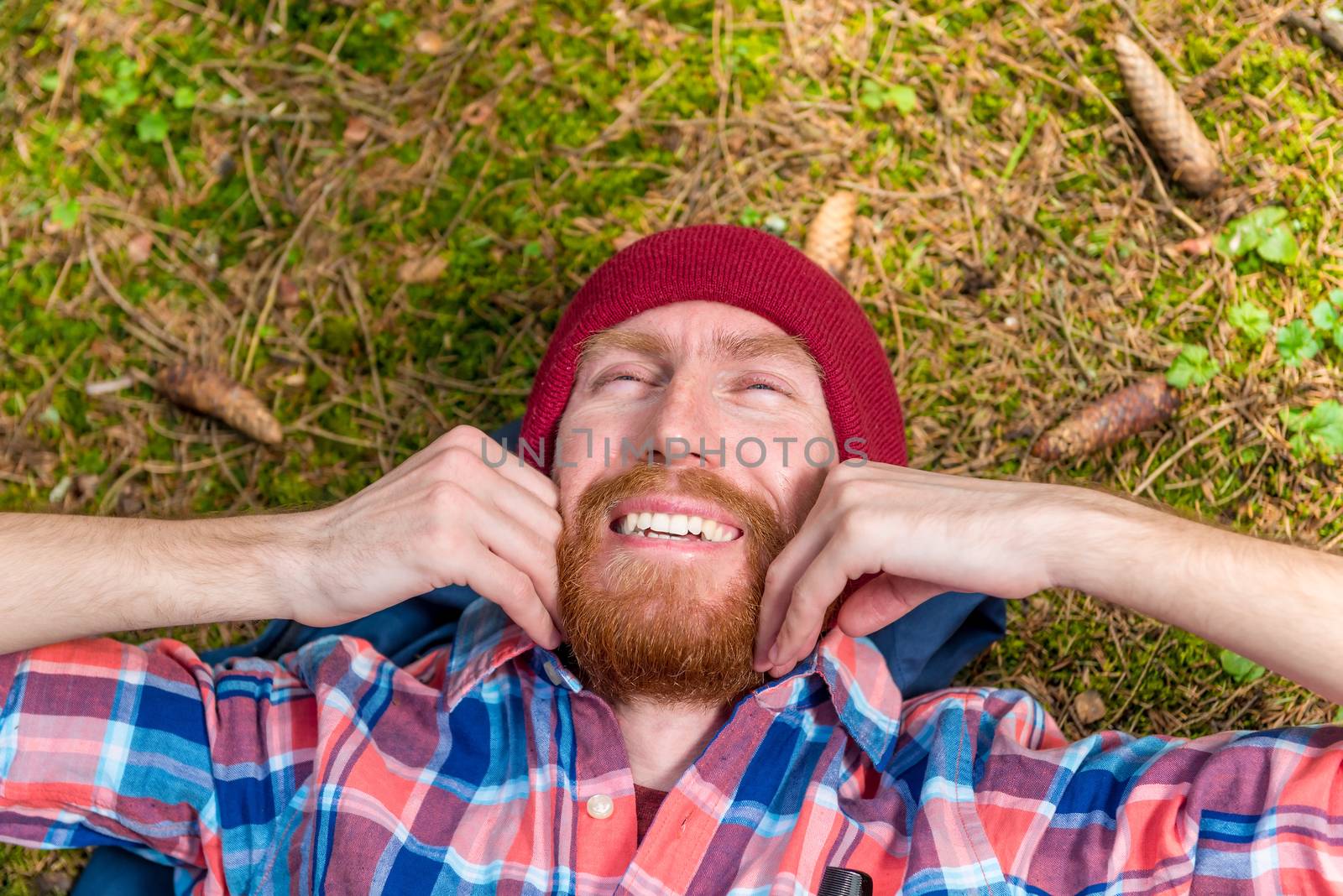 hipster strokes his red beard, a portrait of a curtain, a man lies on the ground in the woods