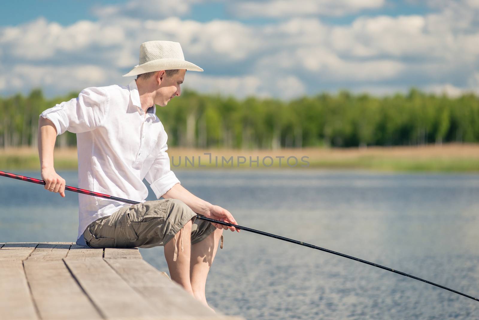 male fisherman with a fishing rod sits on the pier near the lake
