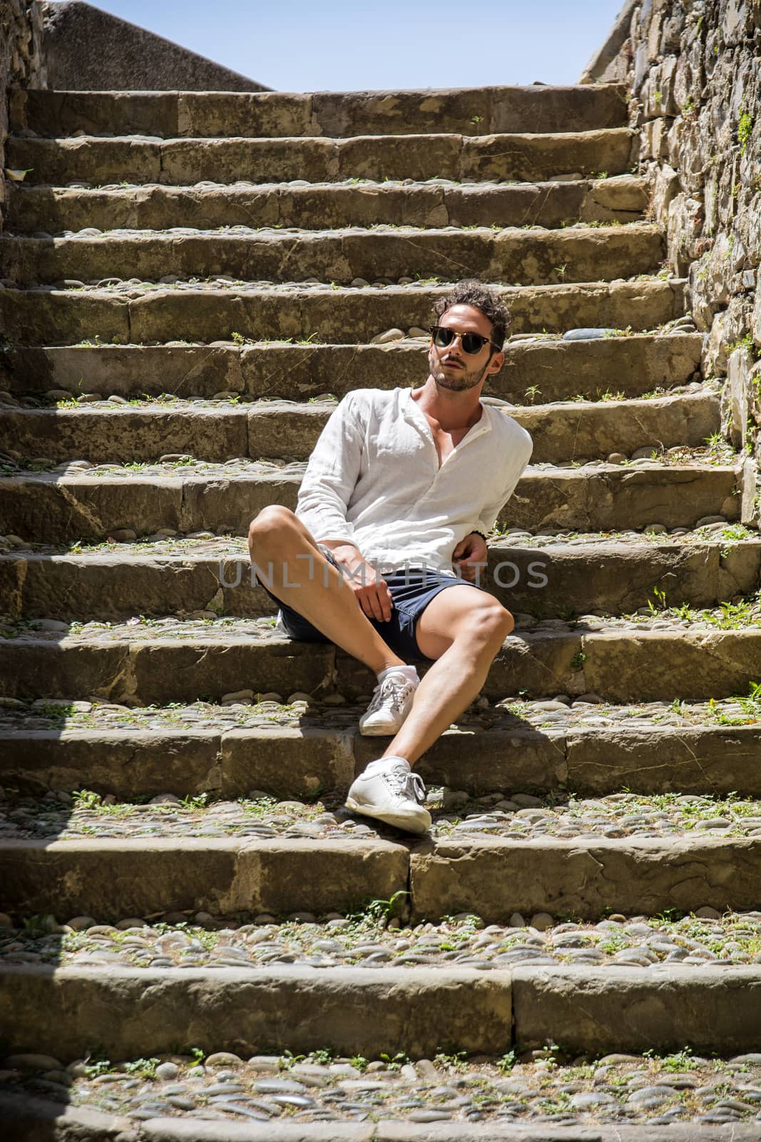 Side view of stylish man in sunglasses sittingon stone stairs and relaxing. Ocean on background.