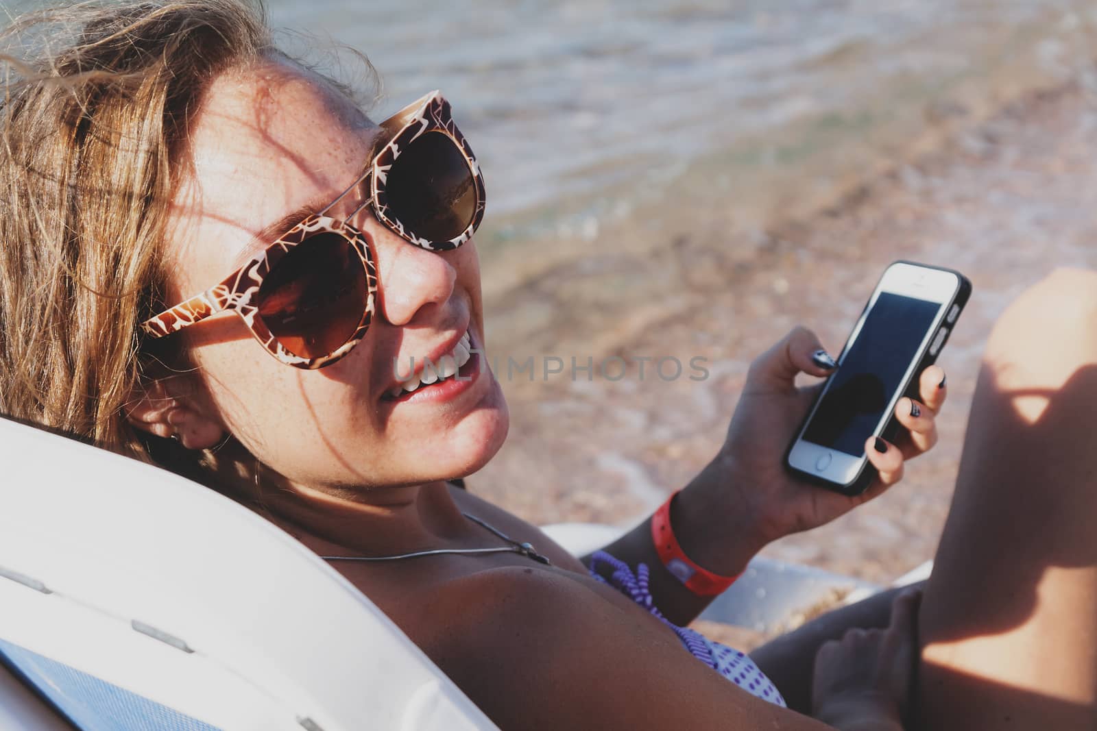 A young tanned woman looks and laughs into a smartphone through sunglasses on a lounger. The concept of a lifestyle is always on the Internet.