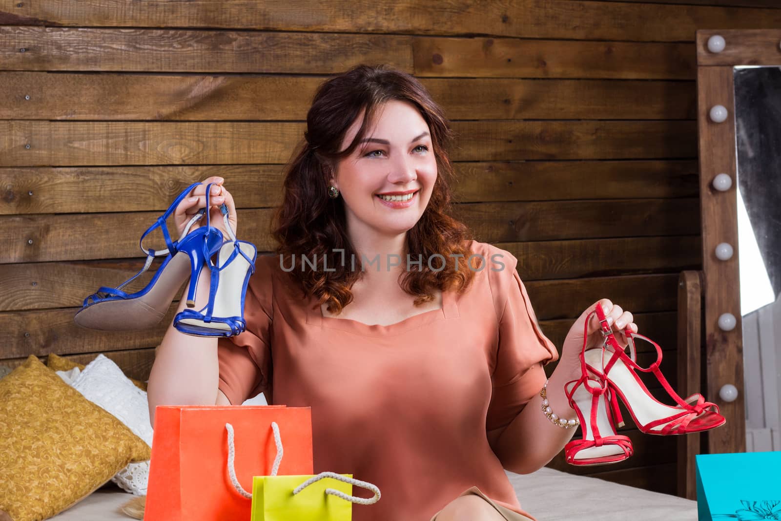 Happy smiling woman after shopping with colourful paper bags and new shoes sits on bed with wooden wall on background