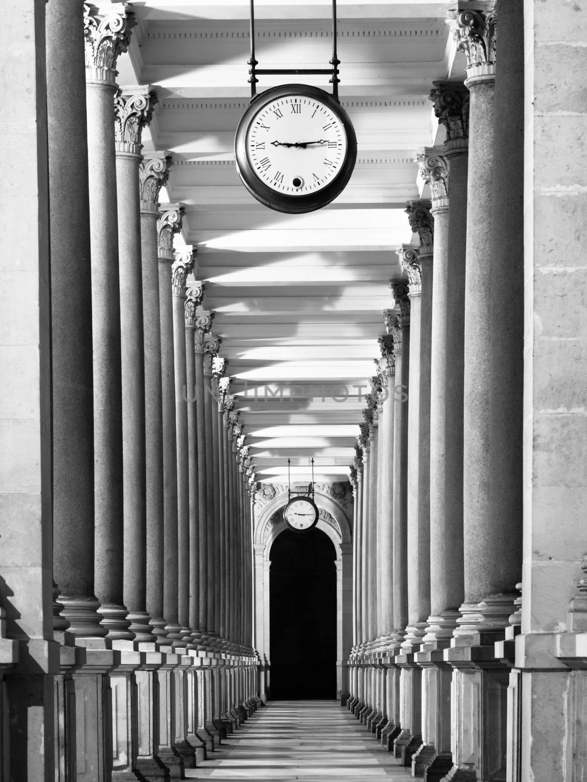 Long colonnafe corridor with columns and clock hanging from ceiling. Cloister perspective. . Black and white image.