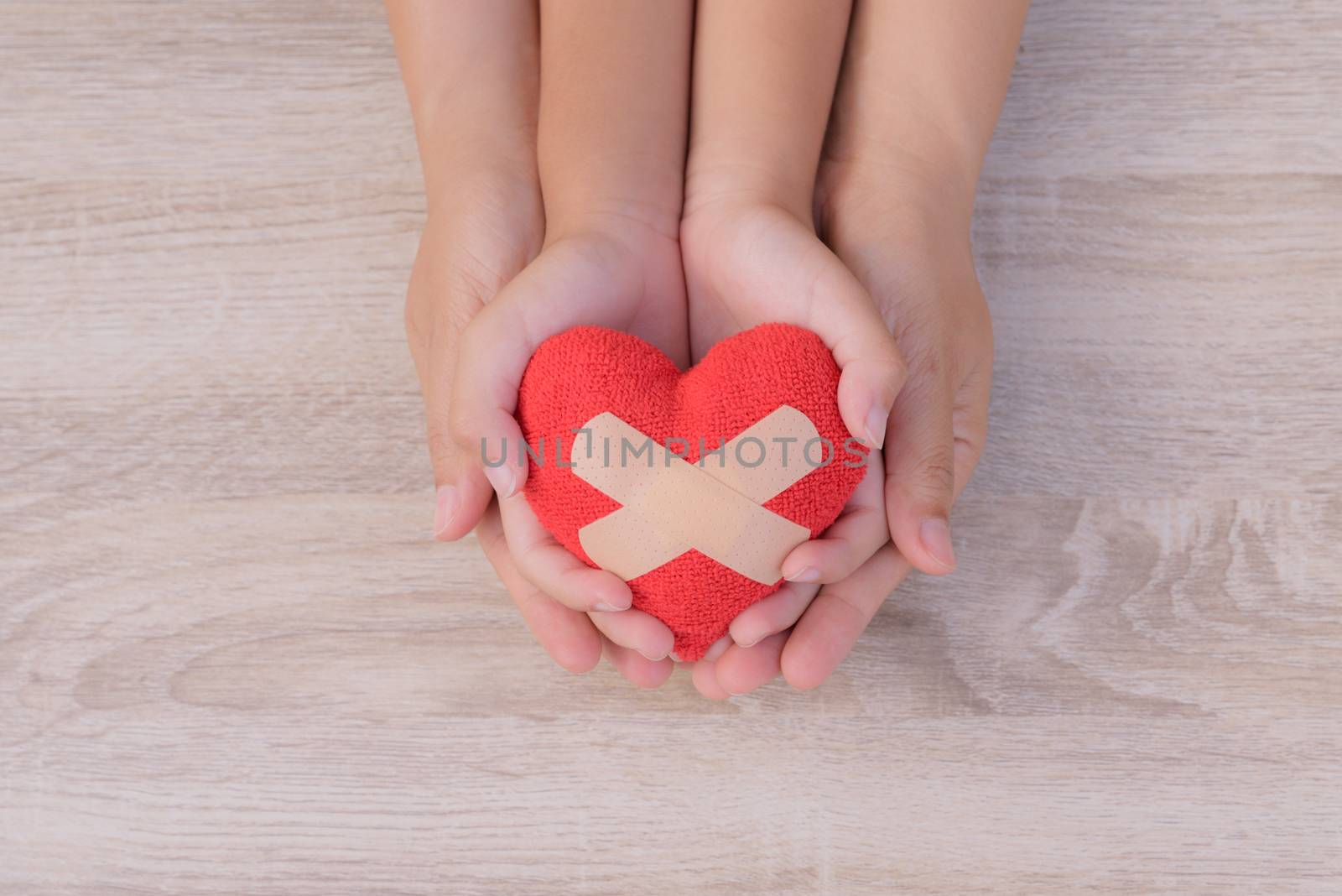 Health care, love, organ donation, family insurance and CSR concept. adult and child hands holding handmade red heart on wooden background. First Aid Band Cushioned Plaster Strip Medical Patch glued on red heart.