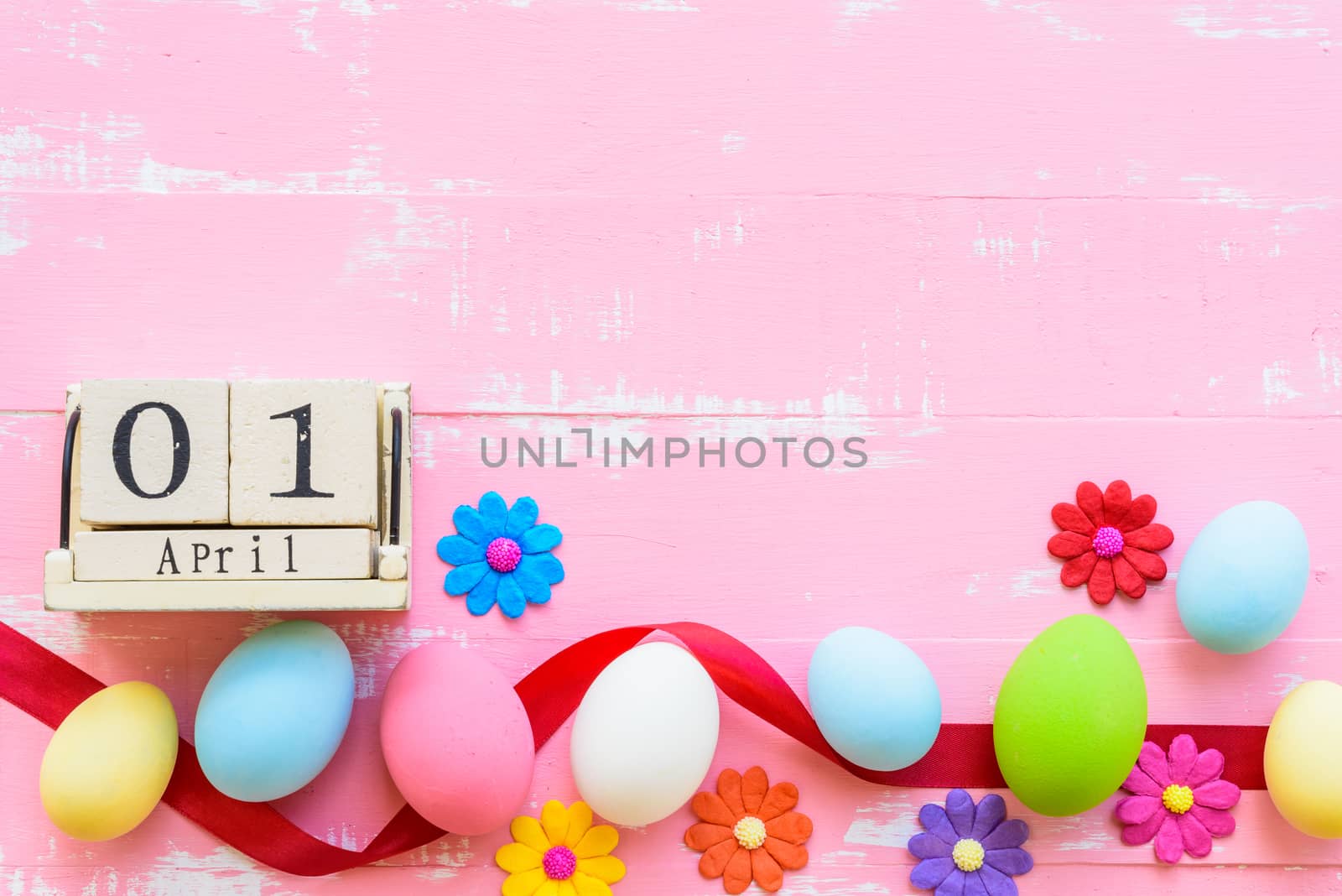 Block wooden calendar for Easter Day, April 1. Row Easter eggs with colorful paper flowers and red ribbon on bright pink and white wooden background.
