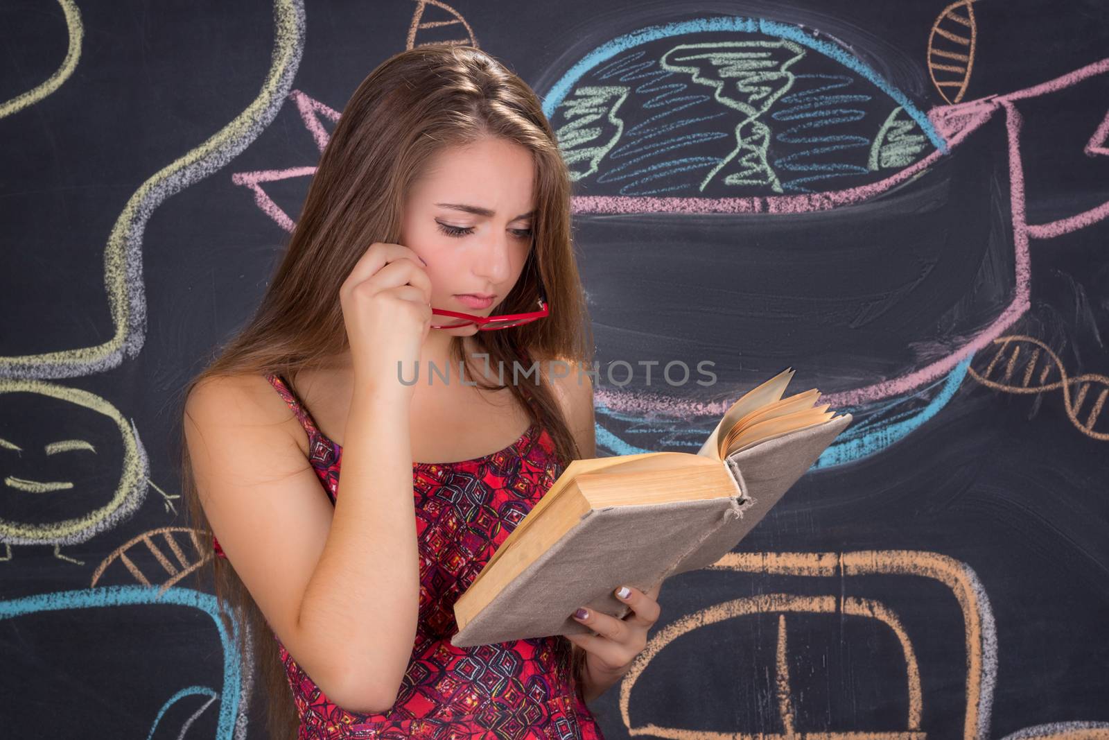 Young student girl in red dress and red glasses reads a book in front of school blackboard