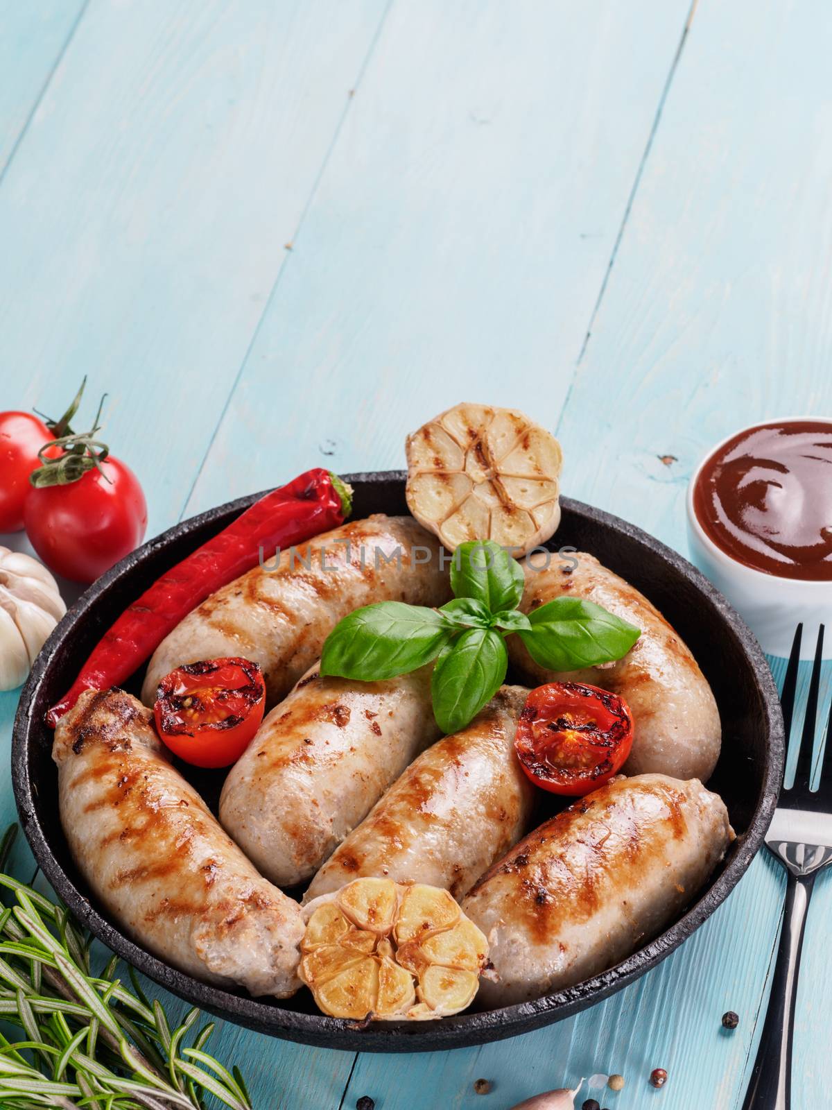 Chicken homemade sausages, sauces ketchup and vegetables and herbs on blue wooden background. Grilled sausages and grilled vegetables in black iron pan. Top view or flat lay. Copy space. Vertical.