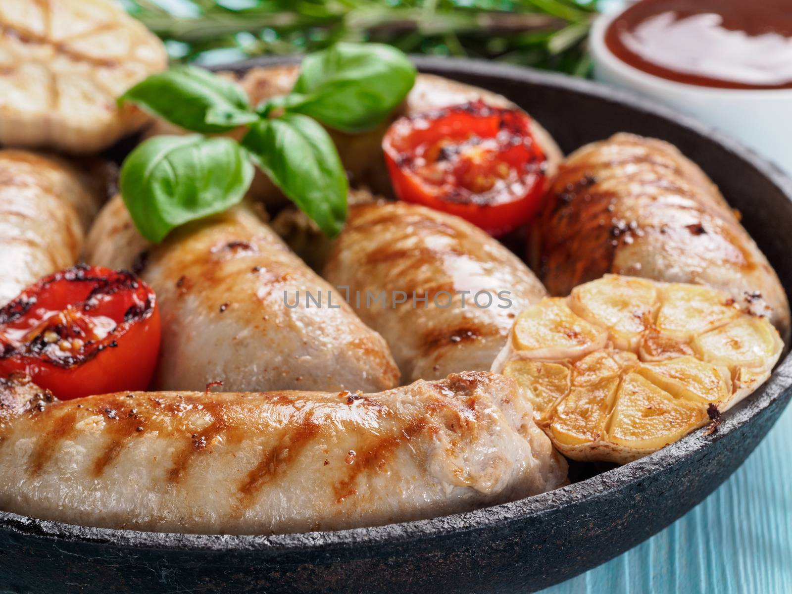 Chicken homemade sausages, sauces ketchup and vegetables and herbs on blue wooden background. Grilled sausages and grilled vegetables in black iron pan