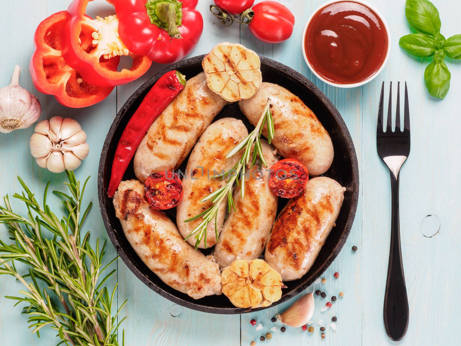 Top view of chicken homemade sausages, sauces ketchup and vegetables and herbs on blue wooden background. Grilled sausages and grilled vegetables in black iron pan. Top view or flat lay.