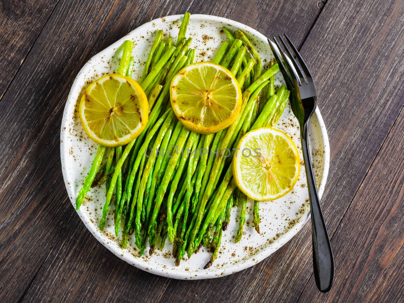 Grilled asparagus and slice of lemon in craft trendy plate on dark brown wooden table. Top view or flat lay.