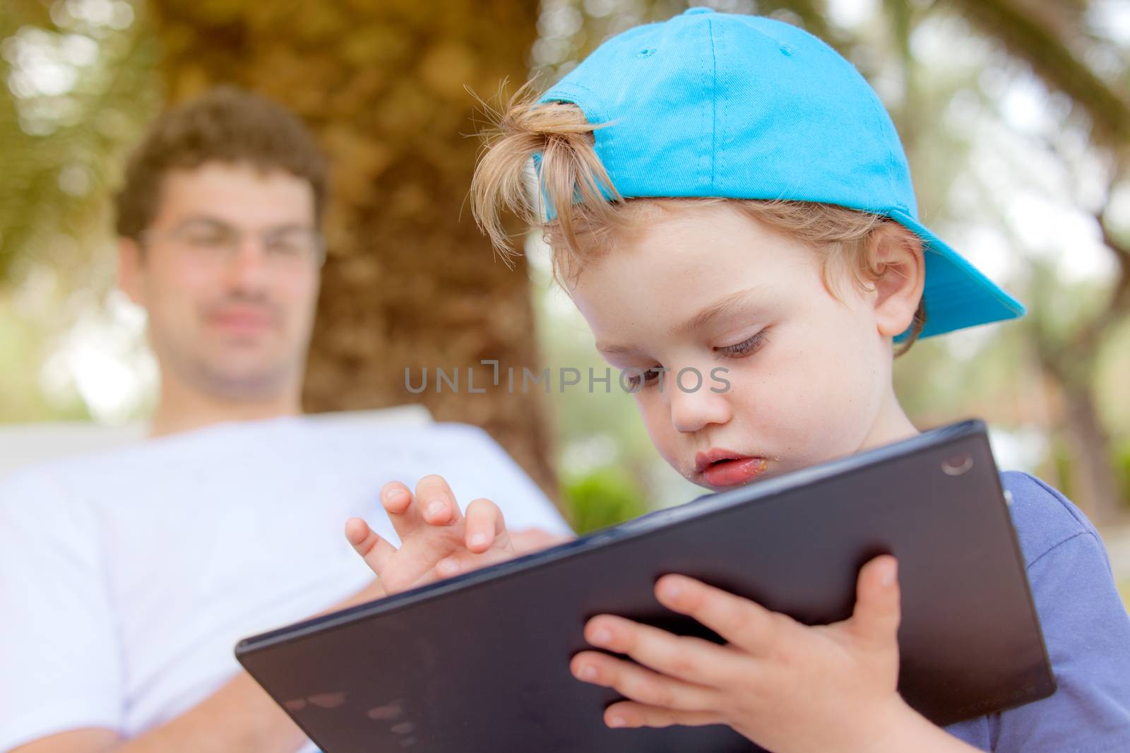 An infant child with food around his mouth is working, playing with tablet outdoors.