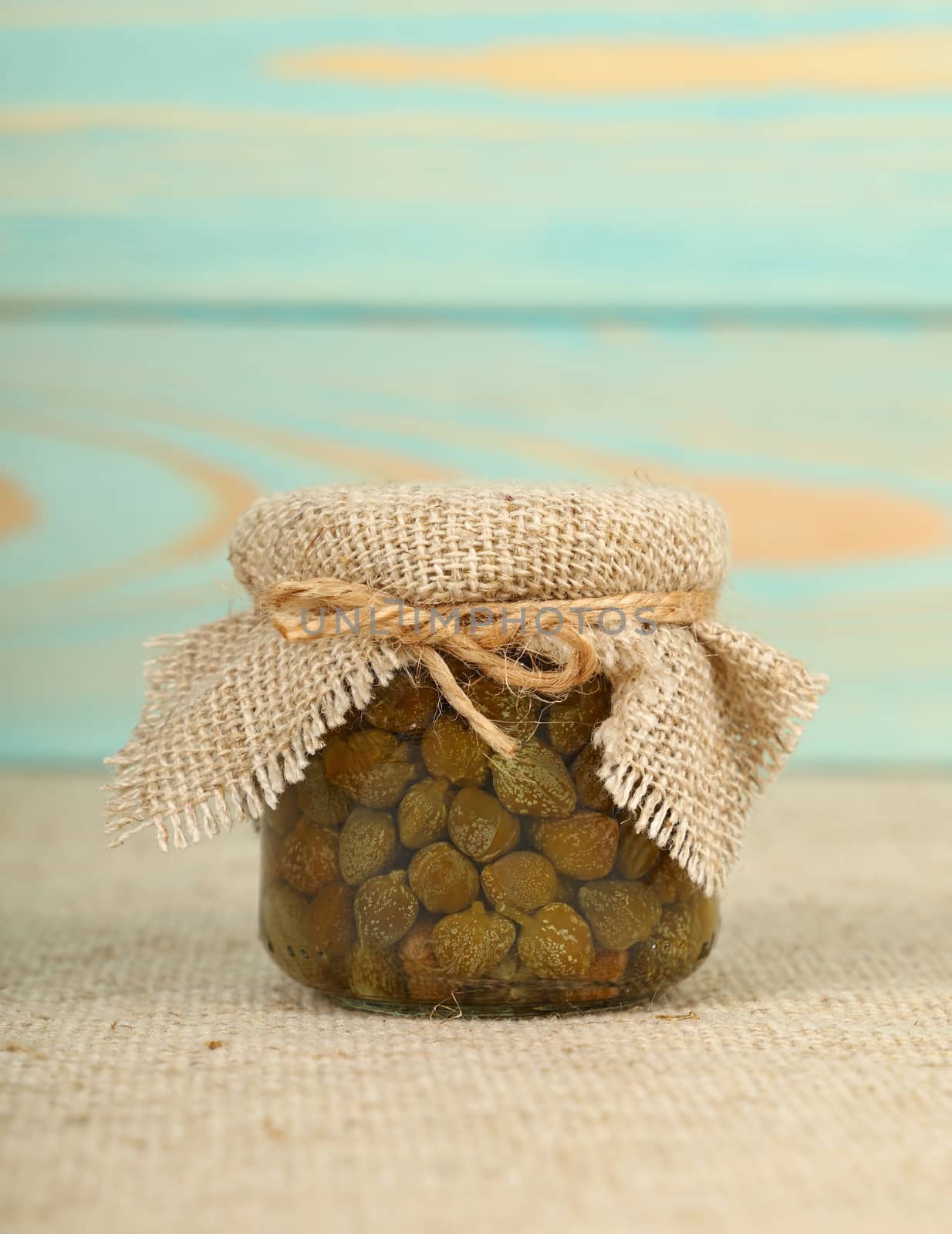 Close up of one small glass jar of pickled capers with canvas top decoration and twine on canvas tablecloth over blue wooden background, low angle side view