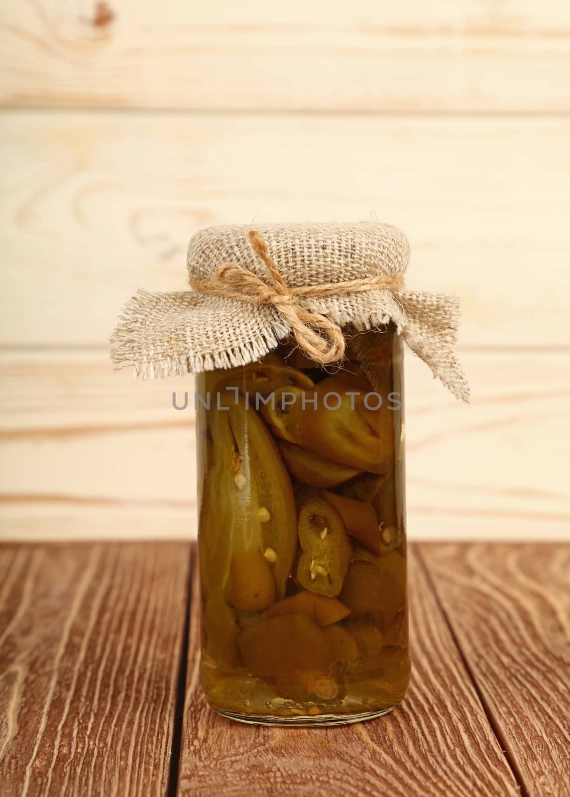 Close up of one small glass jar of pickled cut sliced green hot jalapeno chili peppers with canvas top decoration and twine on brown wooden table over white background, low angle side view