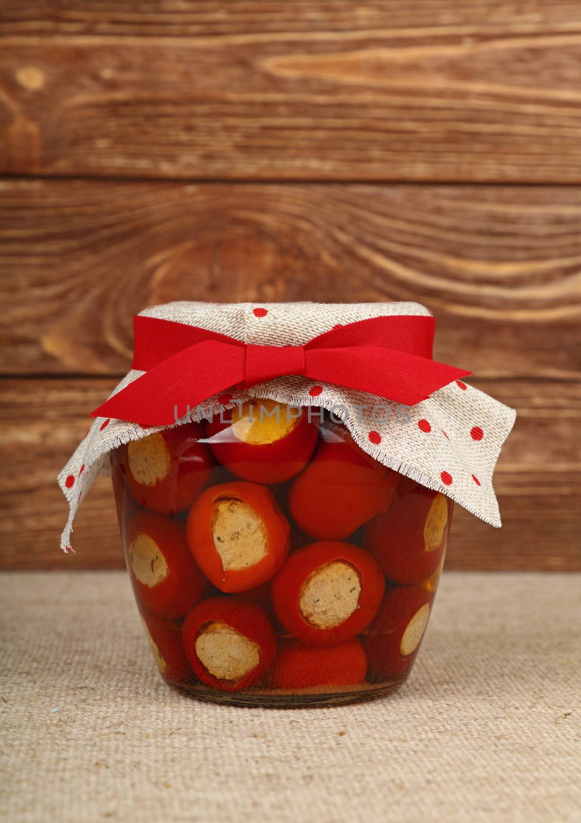 Close up of one glass jar of pickled red hot cherry chili pepperoncini peppers stuffed with soft ricotta cheese with linen canvas lid decoration and red ribbon over brown wooden background, low angle side view