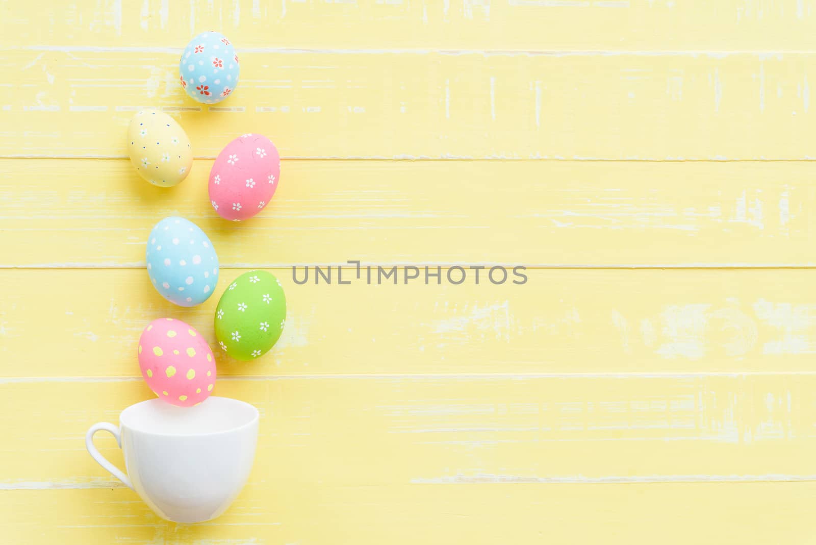 Happy easter! Row colorful Easter eggs spread out from white cup on bright yellow wooden background.