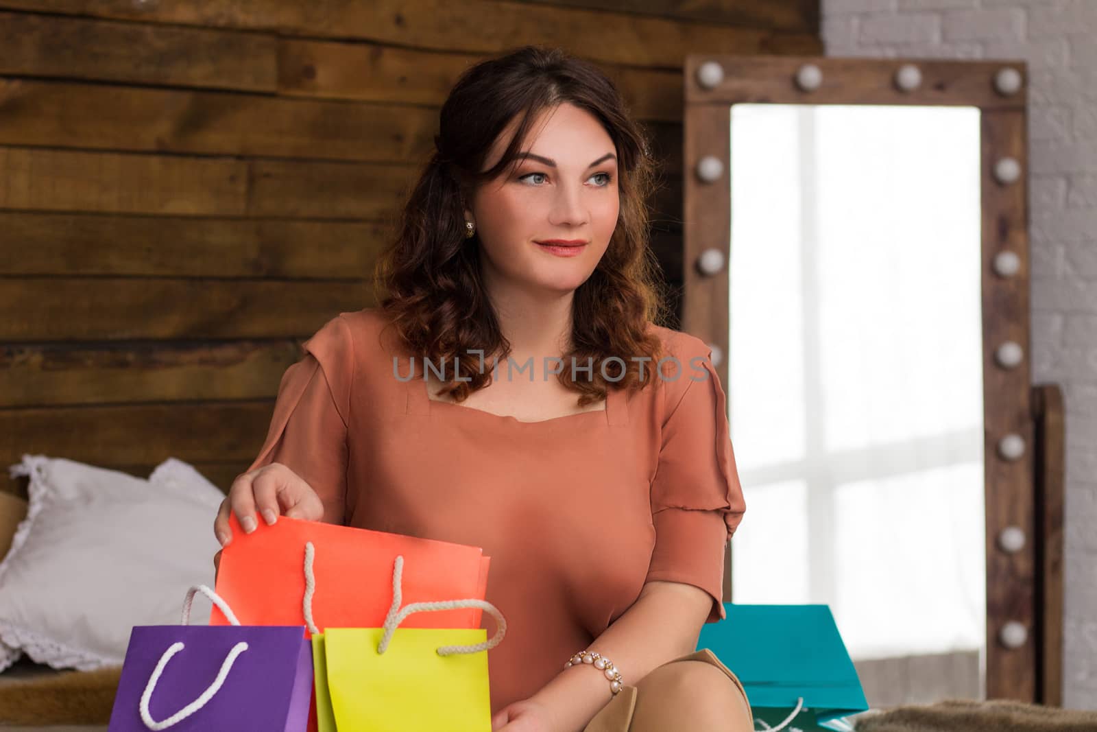 Happy smiling woman after shopping with colourful paper bags sits on bed with wooden wall on background