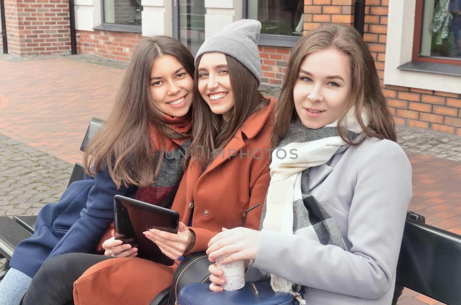 Three girls sit on the bench, holding a tablet and coffee in their hands. Looking at the photographer, smiling
