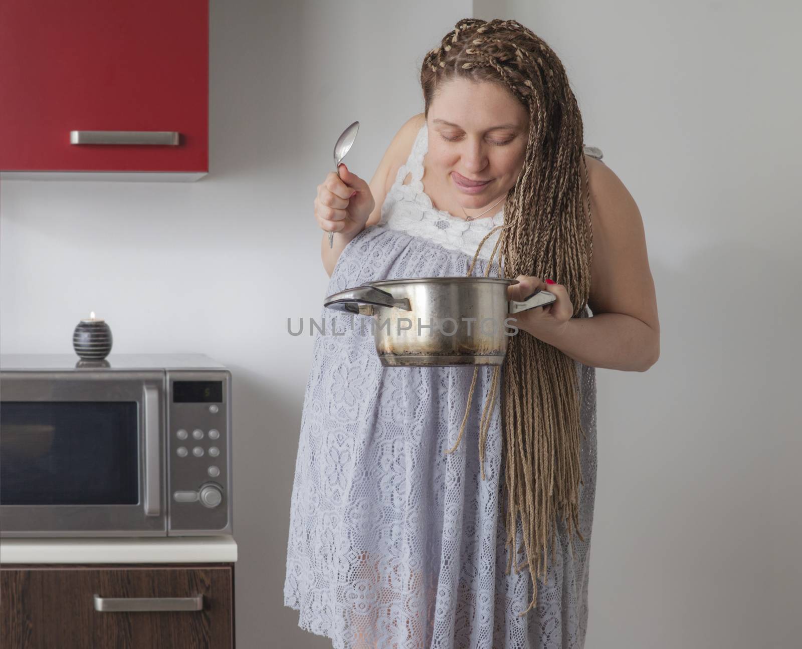 Hungry pregnant woman is holding a saucepan and a spoon, sticking her tounge out.