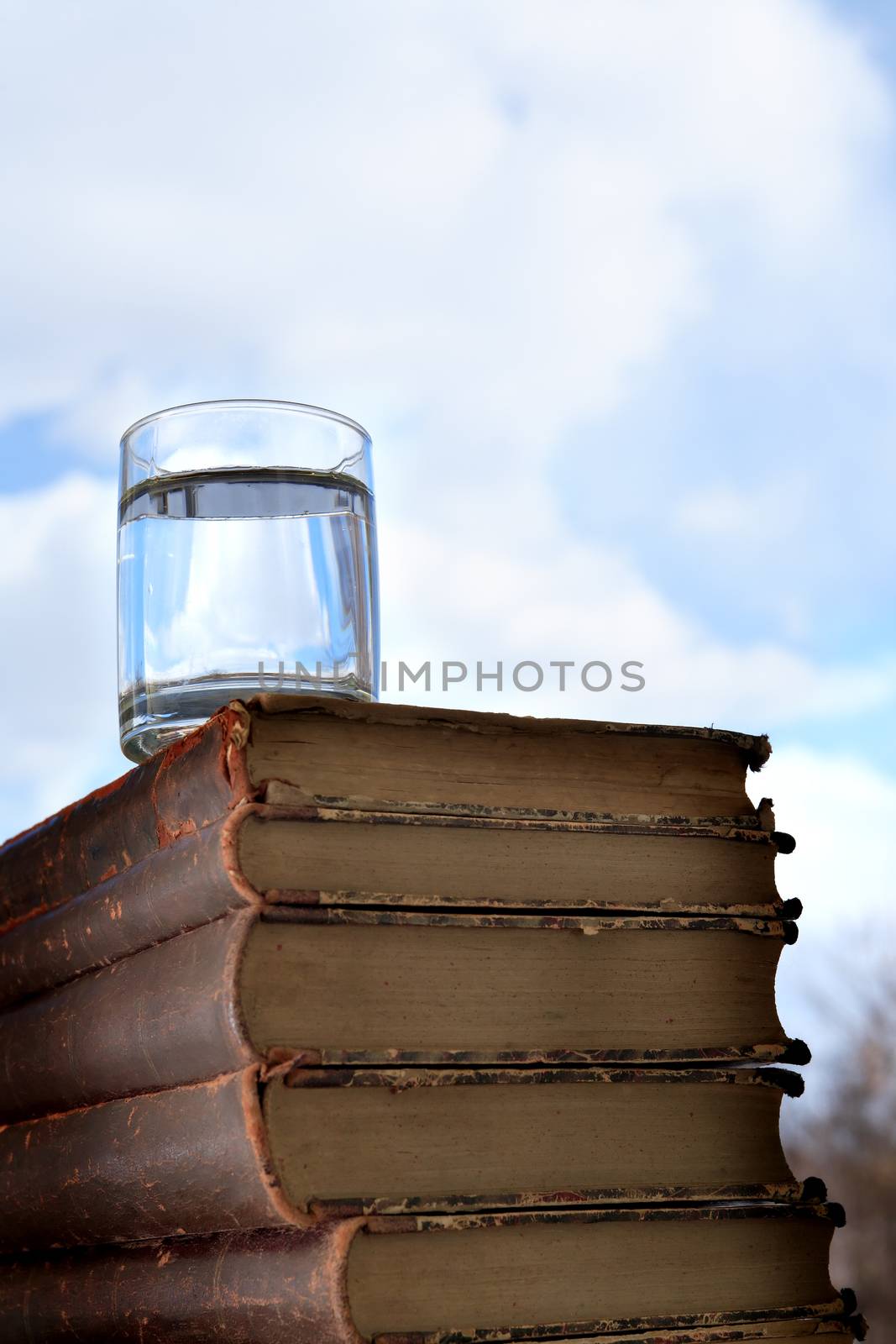 Glass of water on stack of old books on blue sky background with free space
