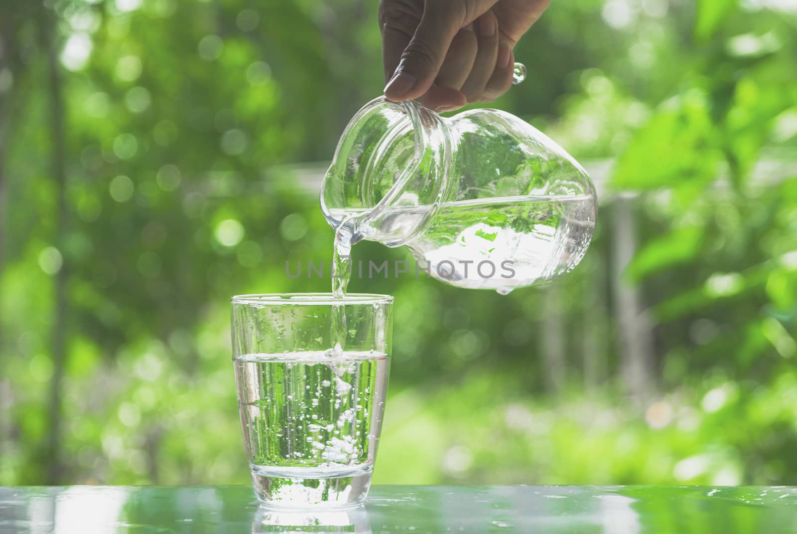 Female hand pouring water from pitcher to glass on nature backgr by kirisa99