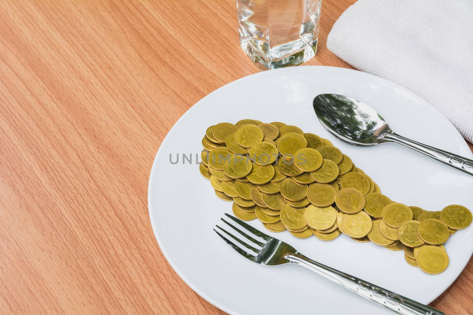 Golden coins shape fish with spoon and fork in white plate and water glass on wooden table.