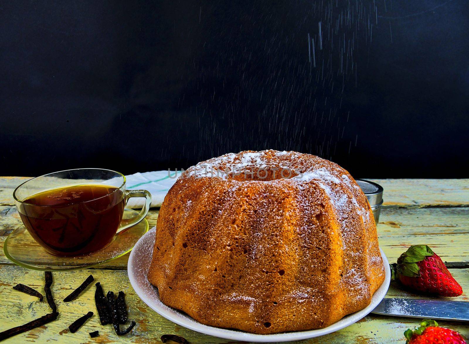 Old fashioned sand cake with cup of black tea and pieces of vanilla on wooden background. Egg-yolk sponge cake on rustic white background. Pouring sugar on sand cake.