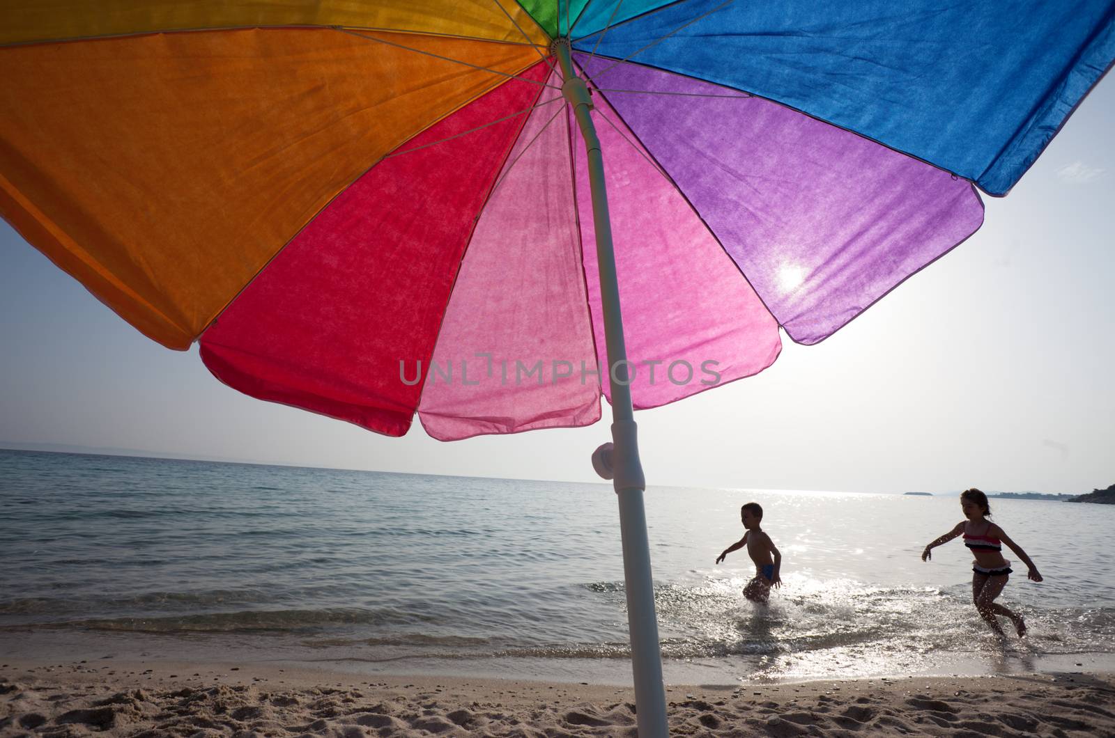 Couple of kids around 6 are playing in the water under the sun, colorful umbrella in foreground.