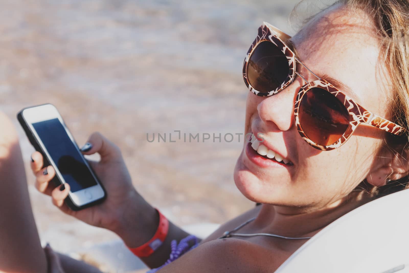 A young tanned woman looks and laughs into a smartphone through sunglasses on a lounger. The concept of a lifestyle is always on the Internet.