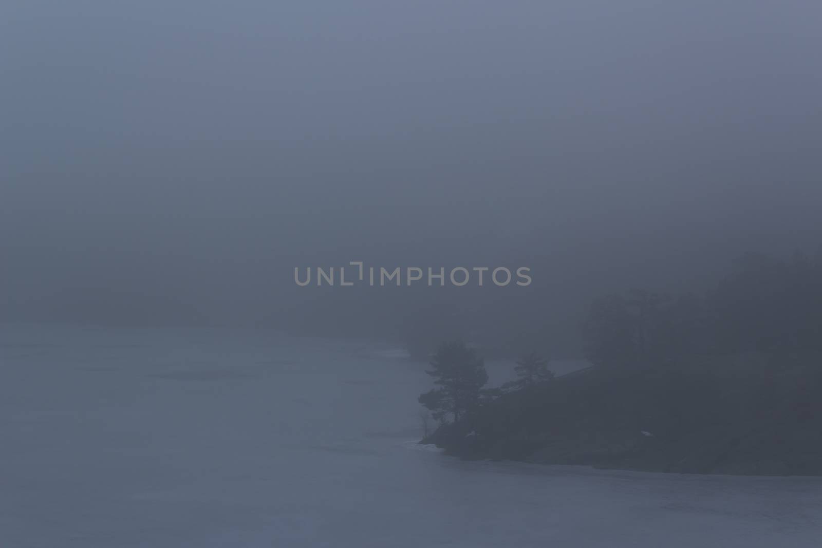 Frozen lake surrounded by small mountains with pine and fir trees on a misty, moody, day. Pine and fir trees on a mountain leading down to a frozen lake on a misty day. Nackareservatet - nature reserve in Sweden