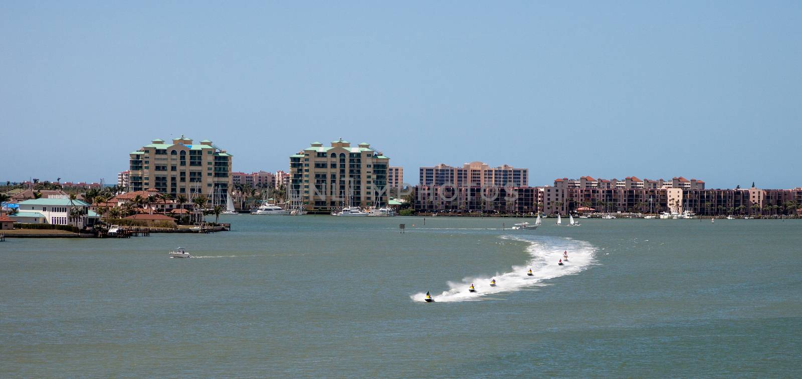 Skyline in the background as a Group of jet skies zip along the ocean in a straight line, making waves in the ocean in the bay in front of Marco Island, Florida