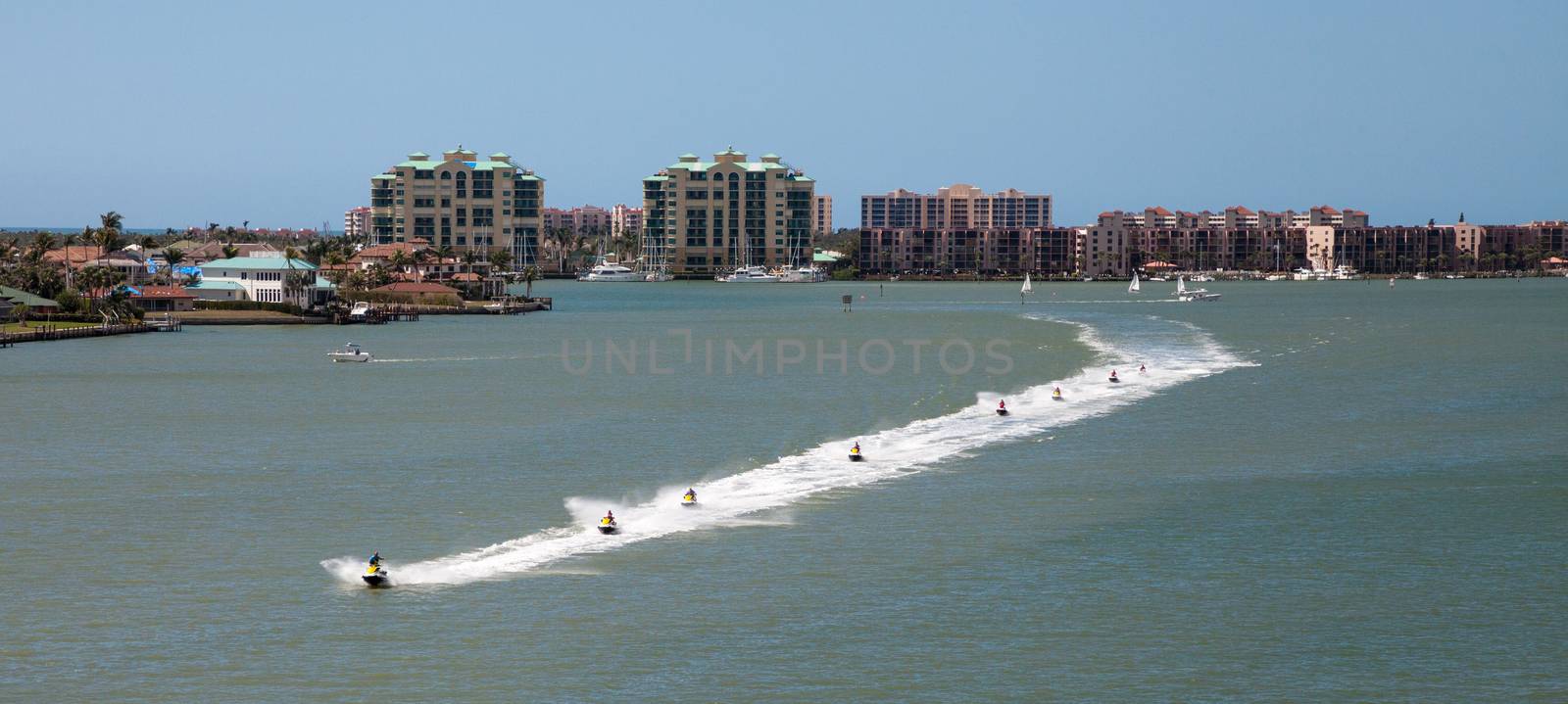 Skyline in the background as a Group of jet skies zip along the ocean in a straight line, making waves in the ocean in the bay in front of Marco Island, Florida
