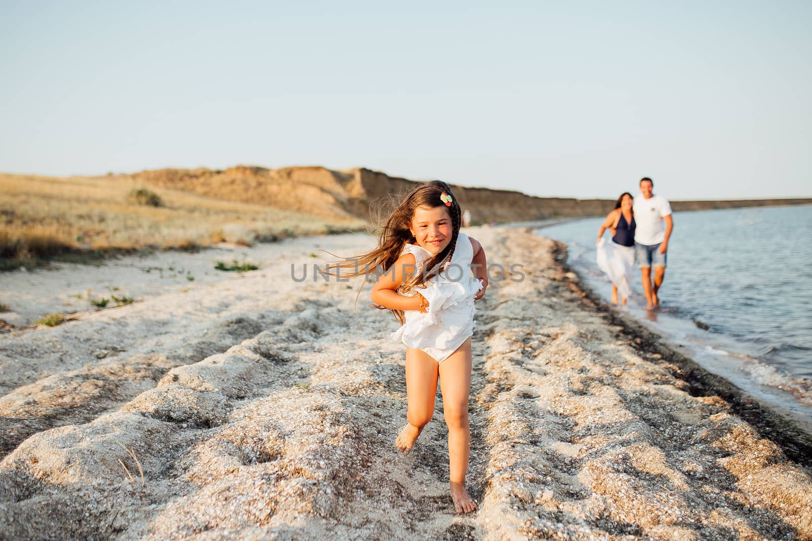 Young happy family walking along the seashore. Little girl with her parents lying on the sand and smiling, parents want to kiss their girl on the cheek.