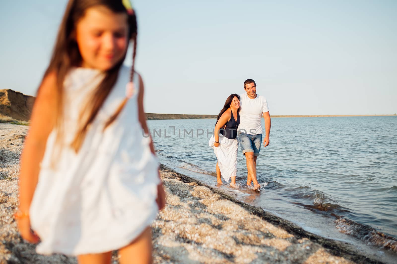 Young happy family walking along the seashore. Little girl with her parents lying on the sand and smiling, parents want to kiss their girl on the cheek.