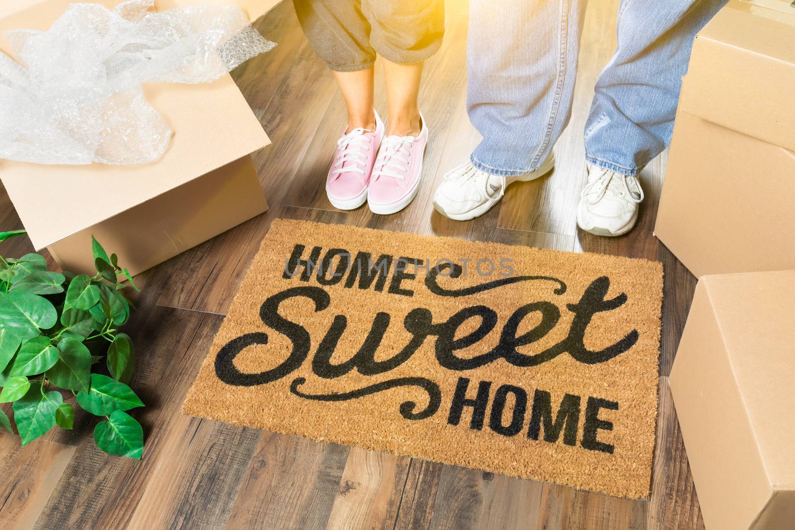 Man and Woman Standing Near Home Sweet Home Welcome Mat, Moving Boxes and Plant.