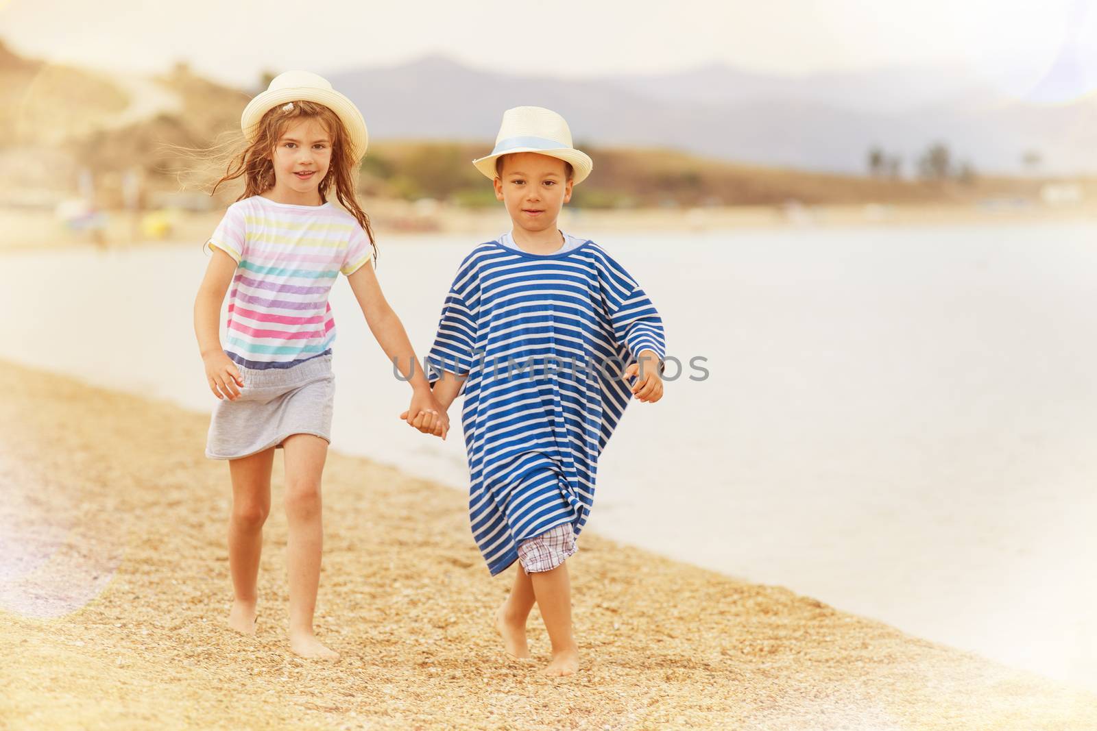 A boy and a girl around six holding hands while taking a walk on the beach smiling.
